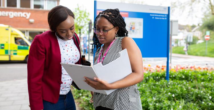 Two young women standing outside a hospital, looking at a clipboard.