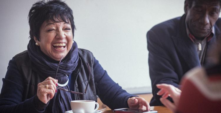 A woman and man sitting with others at a table, smiling and laughing. There are coffee cups on the table.