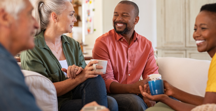 Four adults sitting in a living room, smiling, chatting, and drinking from mugs