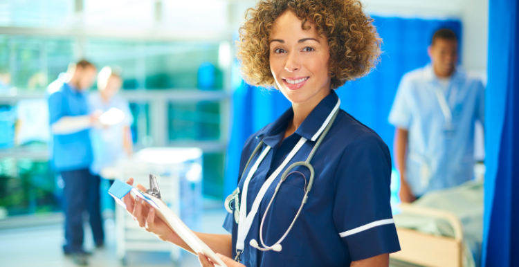 A medical professional standing in a hospital ward, holding a clipboard and wearing a stethoscope 