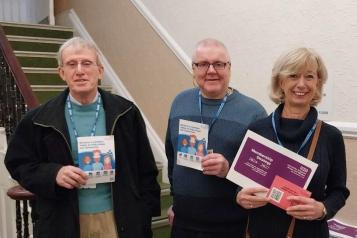 Three people wearing NHS lanyards, smiling and holding information booklets about becoming a UHBW governor.