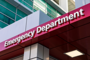 A red sign which reads 'Emergency Department', in front of a hospital