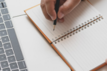 A person writing in a notebook in front of a laptop