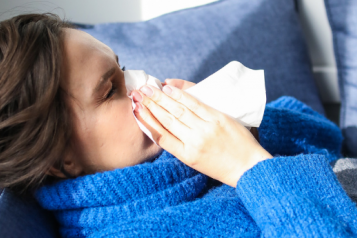 A woman in a blue jumper lying on a sofa and blowing her nose