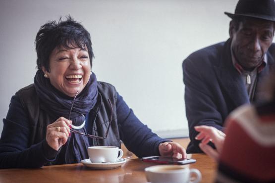 A woman and man sitting with others at a table, smiling and laughing. There are coffee cups on the table.