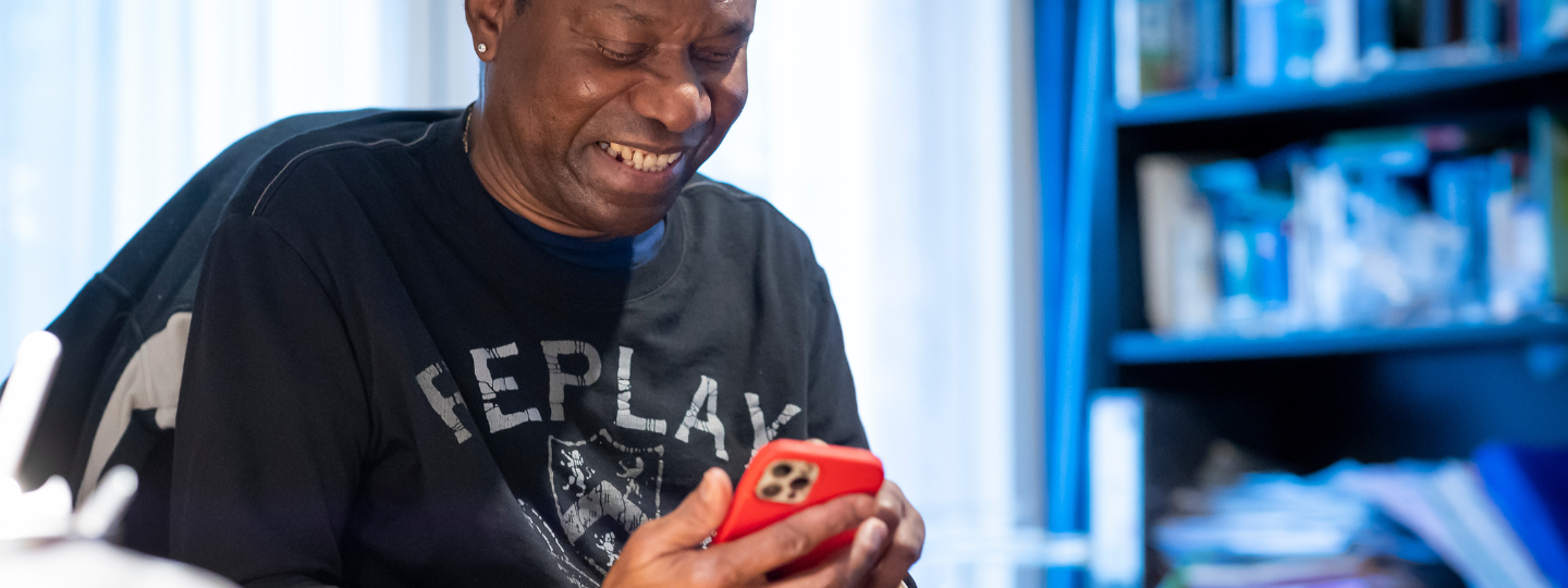 A man sitting at a desk in front of a bookcase, looking down at a mobile phone.